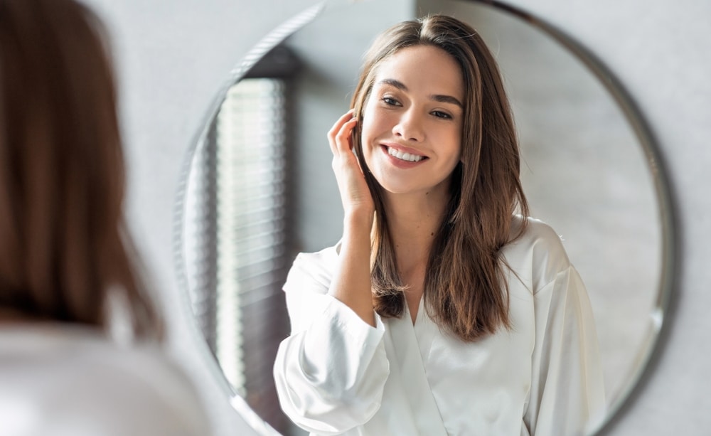 Beautiful woman smiling at herself in the mirror, aging naturally after stopping Botox treatments.